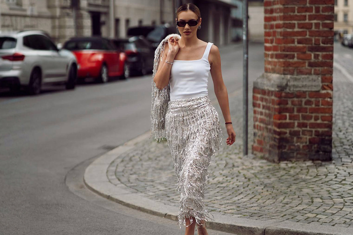 Woman walking on a city street wearing a white tank top and a silver sequin fringe skirt with sunglasses, street style fashion outfit
