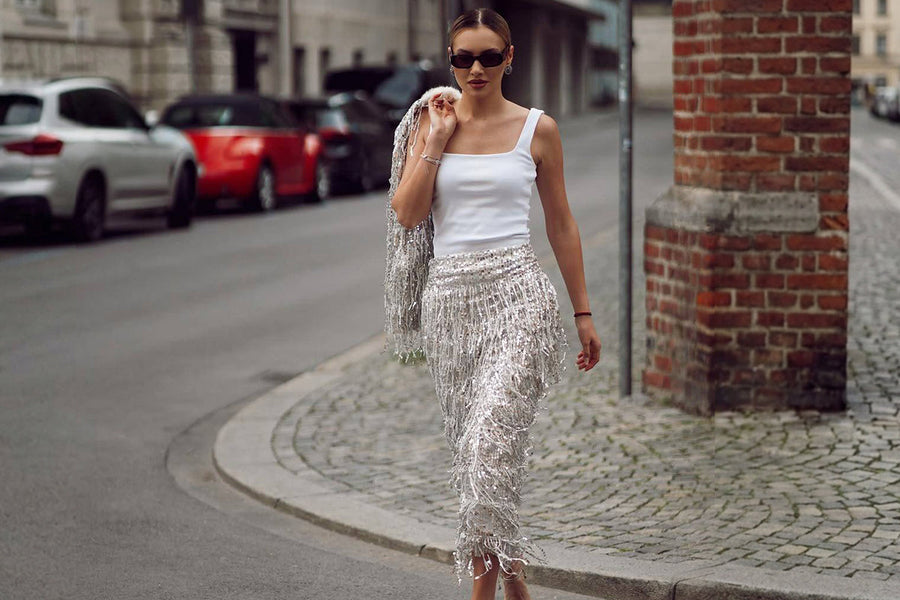 Woman walking on a city street wearing a white tank top and a silver sequin fringe skirt with sunglasses, street style fashion outfit