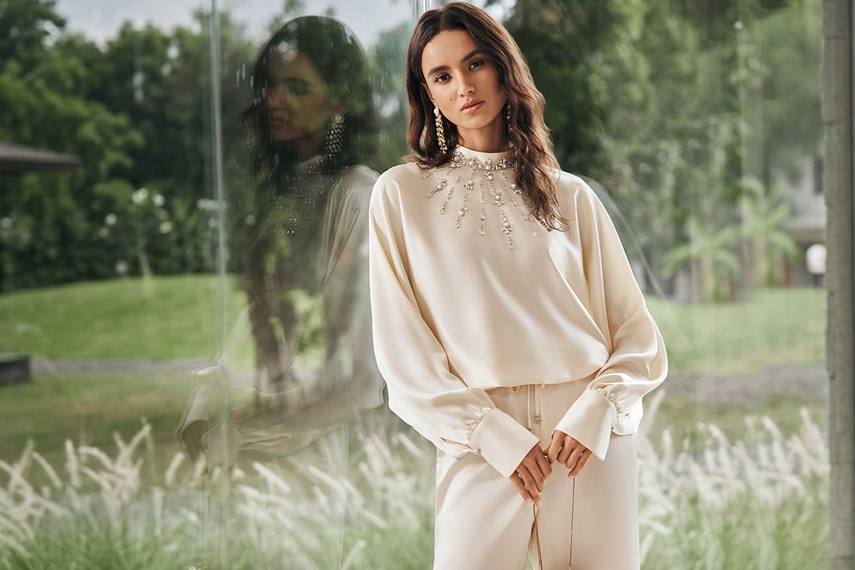 Woman in a cream evening dress with statement earrings and embellished neckline by a glass window.