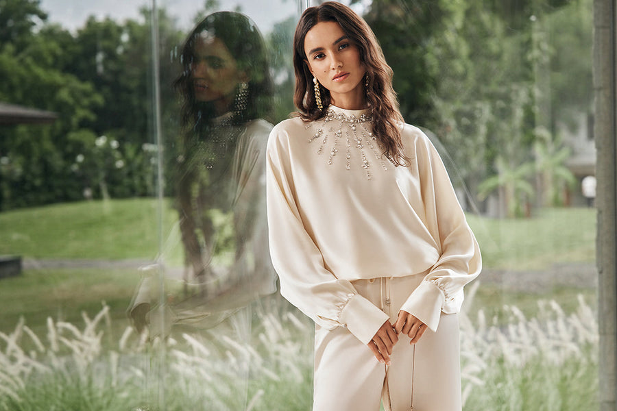 Woman in a cream evening dress with statement earrings and embellished neckline by a glass window.