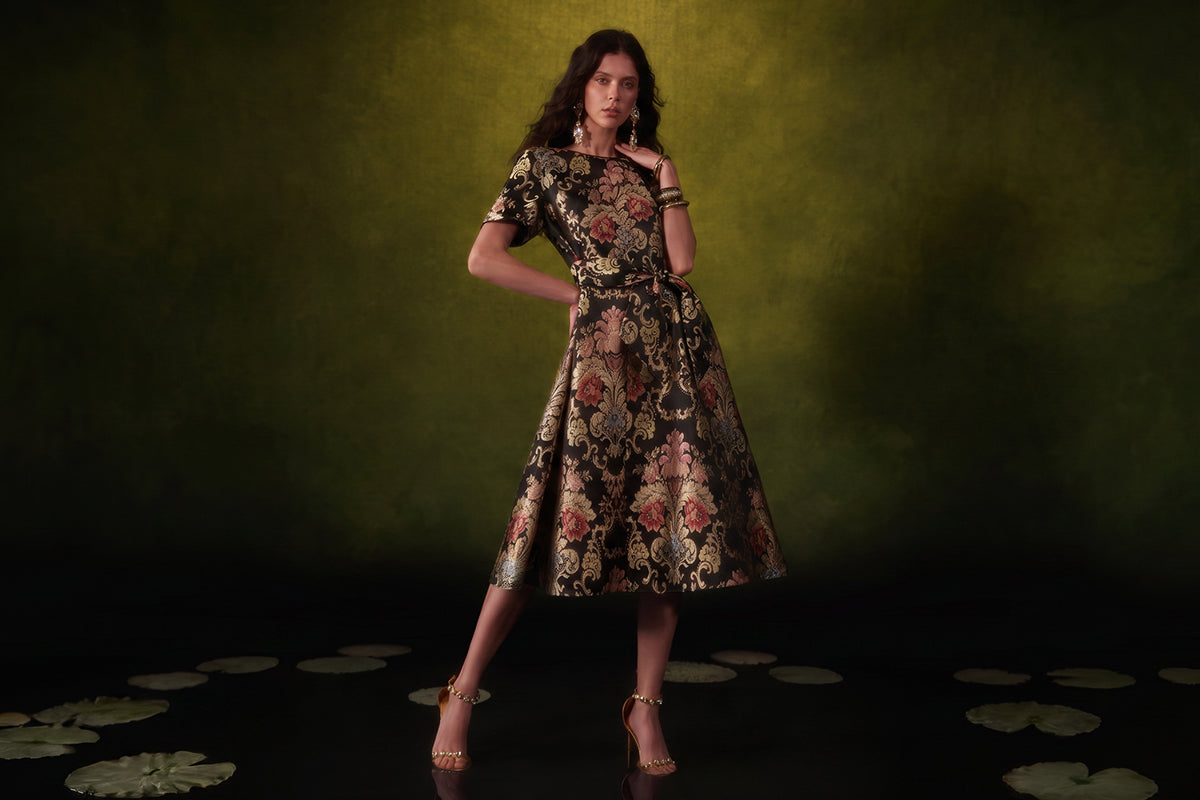 Woman wearing a black and gold floral brocade midi dress with a belted waist and flared skirt